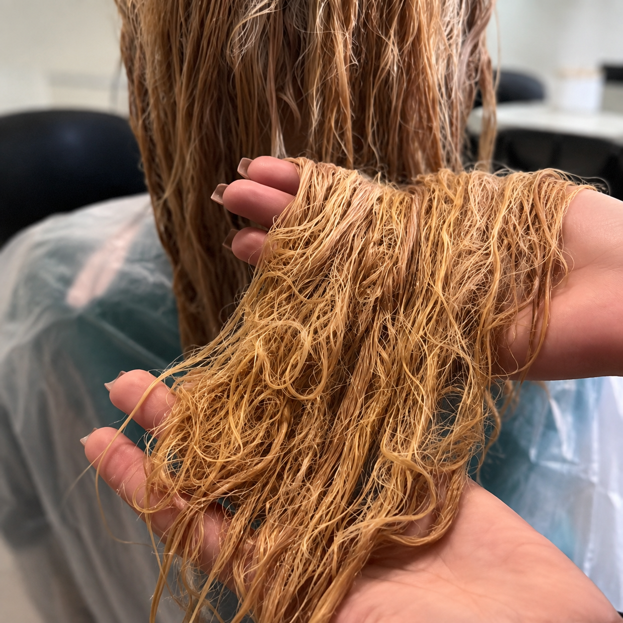 A close-up of wet, tangled light brown hair being held up by two hands, with more of the same hair hanging in the background. The setting appears to be a salon or hair-care environment.